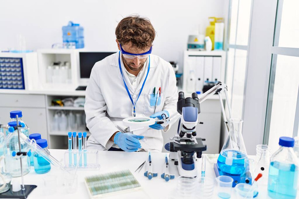 Giovane uomo ispanico indossare uniforme scienziato utilizzando loupe in laboratorio - Foto, immagini