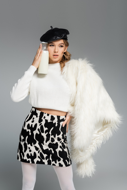 young woman in beret, white faux fur jacket and skirt with cow print holding bottle with milk isolated on grey - Photo, Image