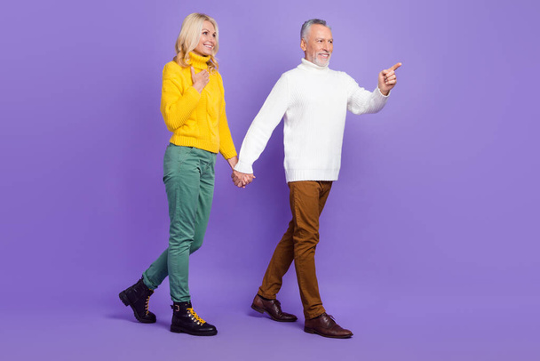 Photo of excited pretty mature wife husband wear knitted pullovers walking holding arms pointing empty space isolated purple color background - Foto, Imagen