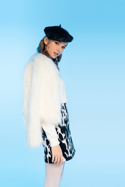 young woman in beret, white faux fur jacket and skirt with cow print looking at camera on blue - Photo, Image