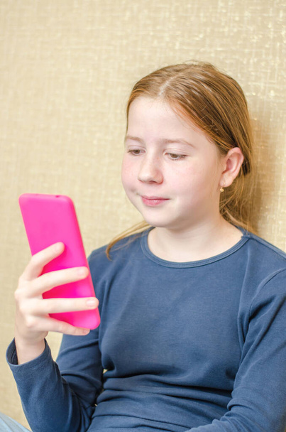 Een meisje van elf kijkt in een smartphone. Het meisje zit op het bed in de kamer en gebruikt de telefoon - Foto, afbeelding