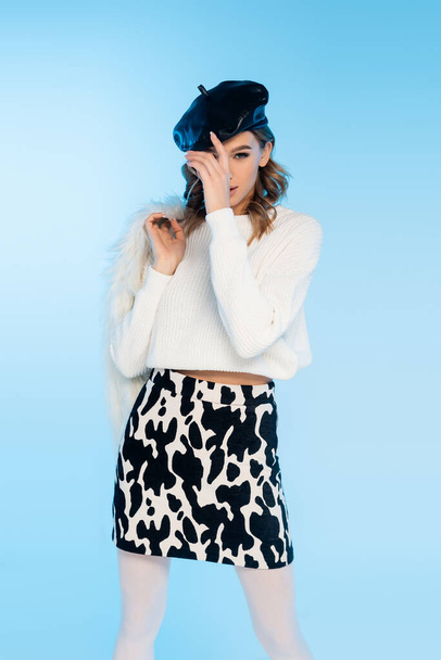 young woman in skirt with cow print holding white faux fur jacket while adjusting beret on blue - Fotoğraf, Görsel