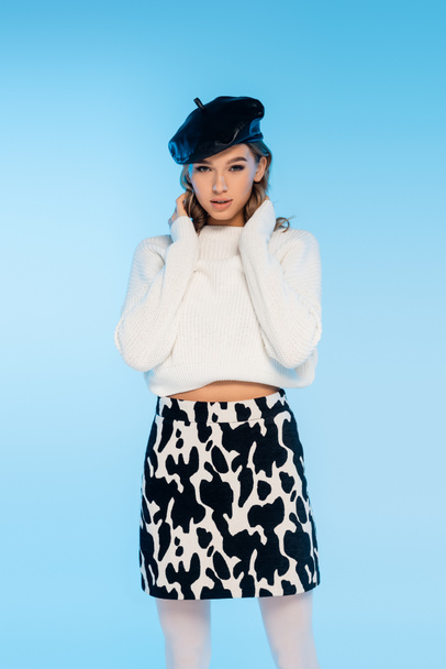 pretty young woman in beret, sweater and skirt with cow print posing on blue - Photo, Image
