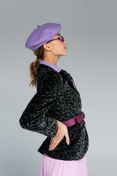side view of stylish woman in beret and blazer with animal print posing with hand on hip isolated on grey  - Foto, imagen