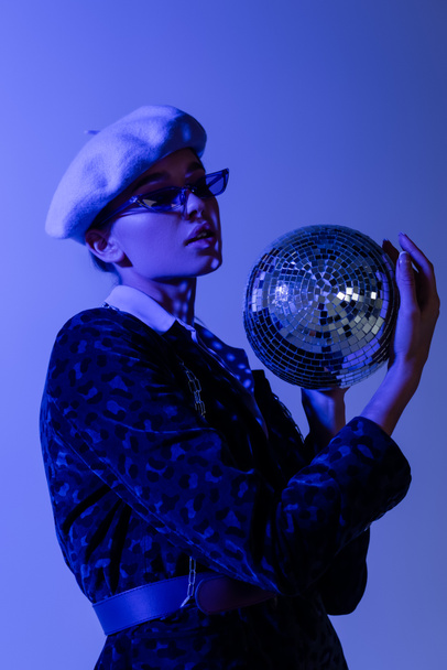 młoda kobieta w berecie, okulary przeciwsłoneczne i marynarka z nadrukiem zwierzęcia gospodarstwa disco ball izolowane na niebiesko  - Zdjęcie, obraz
