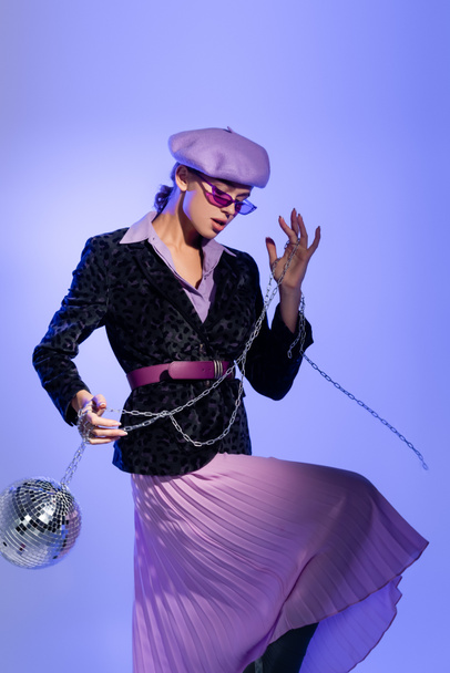 stylowa kobieta w okularach przeciwsłonecznych i marynarce z nadrukiem zwierzęcym trzymająca kulę disco na łańcuszku na fiolecie - Zdjęcie, obraz