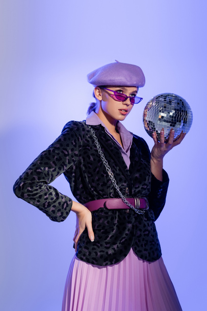 young woman in purple beret and blazer with animal print holding disco ball while posing with hand on hip on violet - Foto, afbeelding