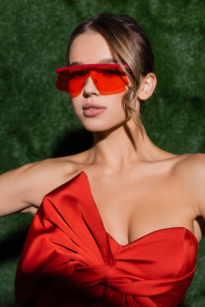 young woman in red blouse and sunglasses looking at camera near grassy background  - Foto, Imagen
