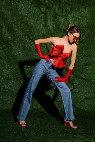 повна довжина жінки в червоній блузці, сонцезахисних окулярах і джинсах позує рукою на стегні біля трав'яного фону
  - Фото, зображення