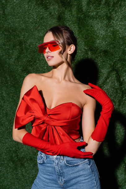 stylish model in red blouse, sunglasses and gloves posing near grassy background  - 写真・画像