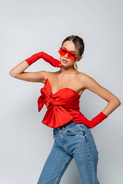 modelo con estilo en blusa roja, gafas de sol y guantes de pie con la mano en la cadera aislado en gris - Foto, imagen