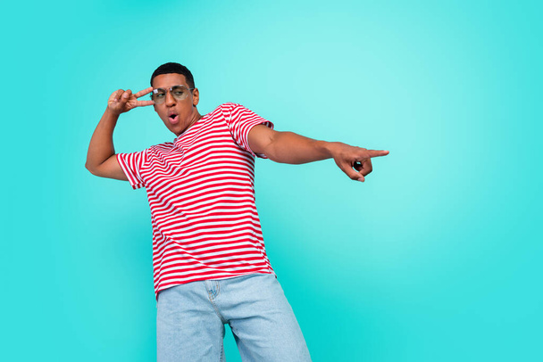 Photo of excited cute young guy dressed red t-shirt dancing pointing empty space showing v-sign isolated turquoise color background - Valokuva, kuva