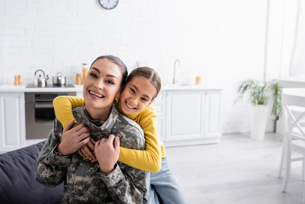Uśmiechnięte dziecko przytulające matkę w mundurze wojskowym w domu  - Zdjęcie, obraz
