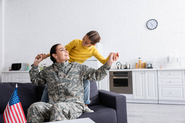 Poika pitää kädestä äitiä sotilaspuvussa lähellä Amerikan lippua kotona.  - Valokuva, kuva