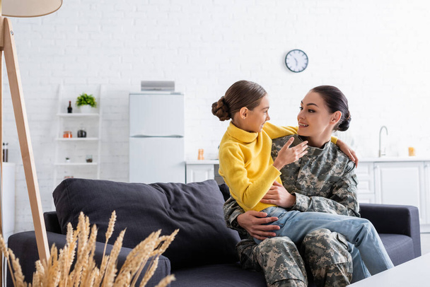Femme souriante en uniforme de camouflage étreignant sa fille sur le canapé à la maison  - Photo, image