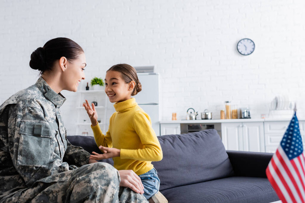 Smiling kid talking to mother in military uniform near blurred american flag at home  - Фото, изображение