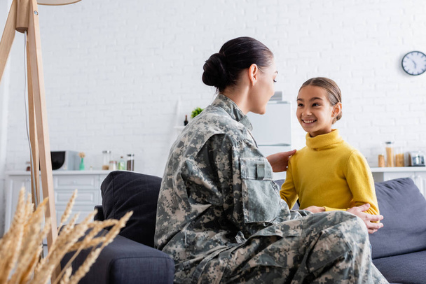 Mulher de camuflagem uniforme olhando para a filha no sofá em casa  - Foto, Imagem