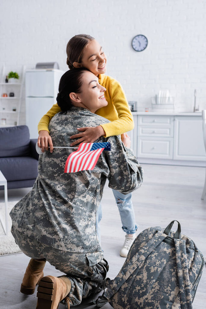 Kid with closed eyes and american flag hugging mother in military uniform near backpack at home  - Photo, Image