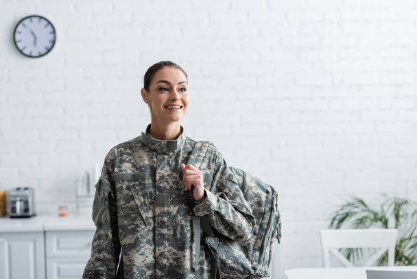 Cheerful woman in military uniform holding backpack at home  - Фото, изображение