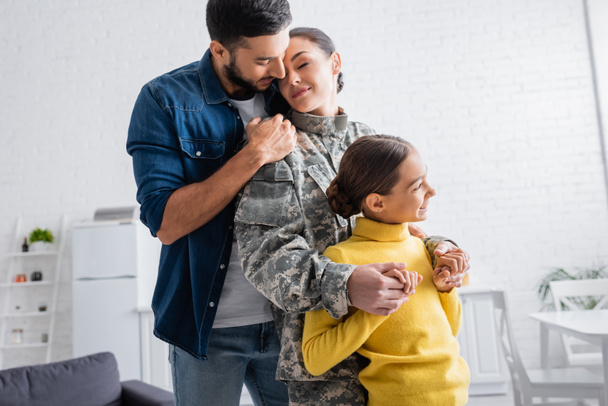 Χαμογελώντας παιδί κρατώντας τα χέρια της μητέρας σε στρατιωτική στολή κοντά στον μπαμπά στο σπίτι  - Φωτογραφία, εικόνα