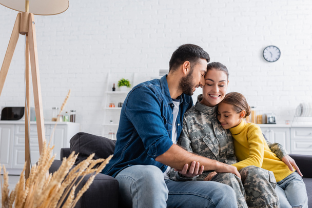 Smiling soldier in uniform sitting near child and husband on couch at home  - Foto, imagen