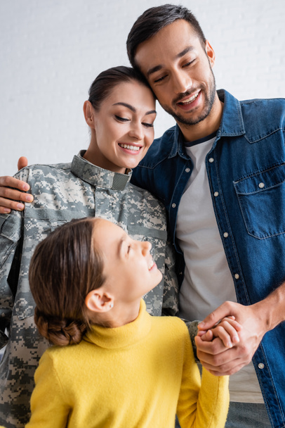 Smiling man holding hand of child and hugging wife in military uniform at home  - Photo, Image