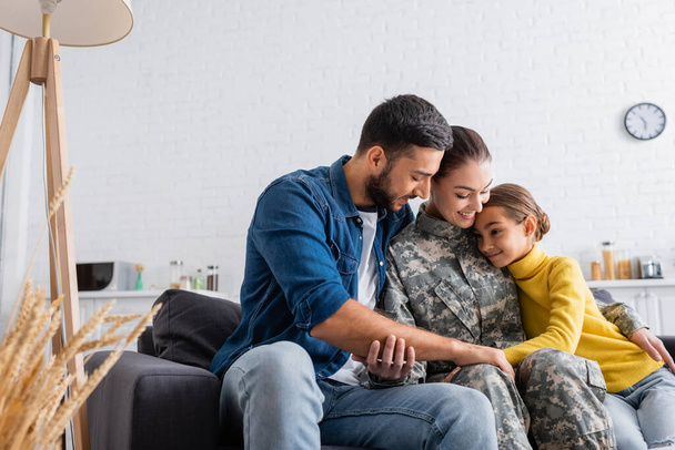 Positive woman in military uniform sitting near child and husband on couch at home  - 写真・画像