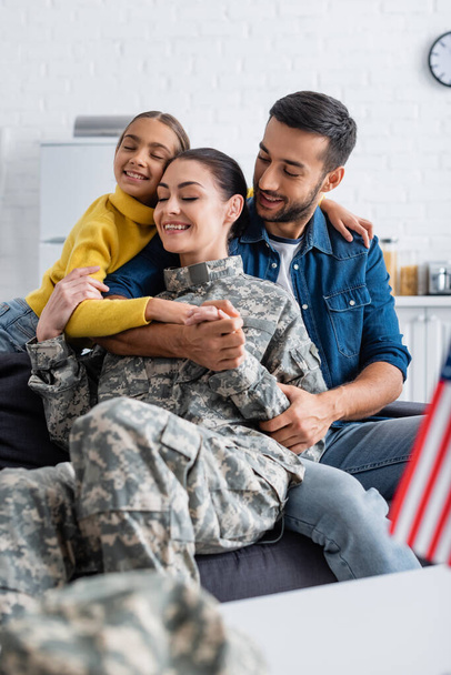 Enfant souriant embrassant la mère en uniforme militaire près du père et drapeau américain flou à la maison  - Photo, image