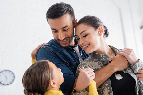 Homem positivo abraçando esposa em uniforme militar perto de filha pré-adolescente em casa  - Foto, Imagem
