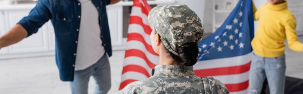 Vrouw in militair uniform staat in de buurt wazig familie met Amerikaanse vlag thuis, banner  - Foto, afbeelding