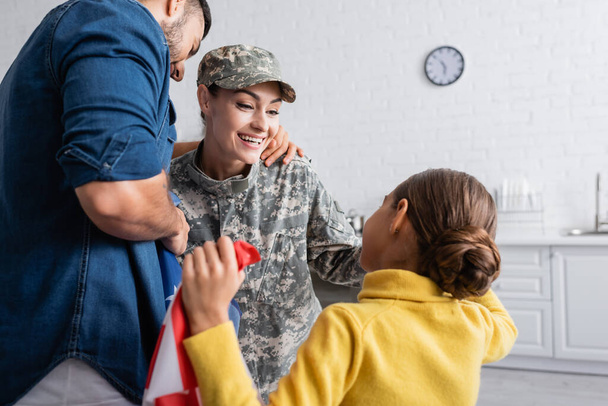 Usmívající se žena ve vojenské uniformě a čepice při pohledu na dceru poblíž manžela s americkou vlajkou doma  - Fotografie, Obrázek