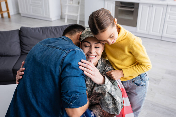 Woman with closed eyes in military uniform embracing husband near kid with american flag at home  - Fotografie, Obrázek