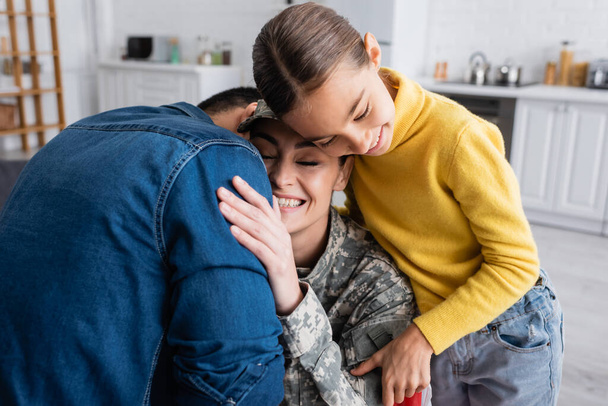 Улыбающаяся женщина в военной форме обнимает мужа рядом с дочерью-подростком дома  - Фото, изображение