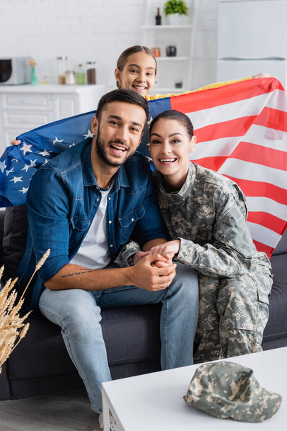Happy woman in military uniform holding hands of husband near smiling kid with american flag looking at camera at home  - 写真・画像