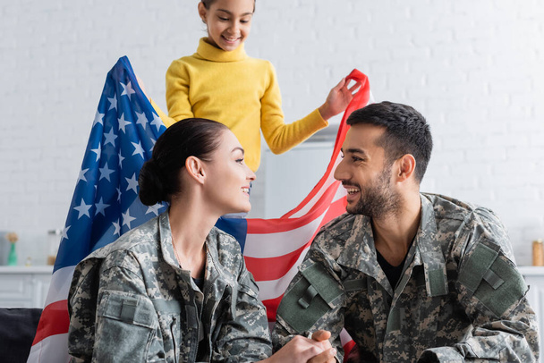 Vrolijke ouders in militair uniform kijken elkaar in de buurt dochter met Amerikaanse vlag thuis  - Foto, afbeelding