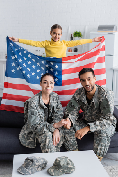 Criança feliz segurando bandeira americana perto dos pais em uniforme militar no sofá em casa  - Foto, Imagem
