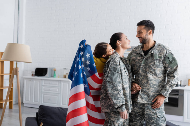 Lapsi, jolla on Amerikan lippu lähellä hymyileviä vanhempia sotilaspuvussa kotona  - Valokuva, kuva