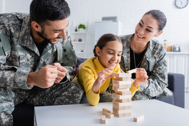 Glückliche Eltern in Militäruniform zeigen Ja-Geste bei Holzklötzchen-Spiel zu Hause  - Foto, Bild