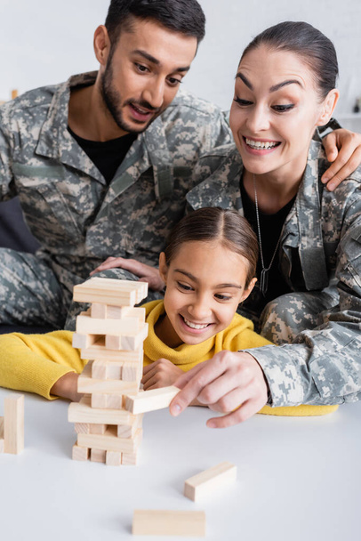 Fröhliche Frau in Militäruniform zeigt auf Holzklötze neben Mann und Kind zu Hause  - Foto, Bild