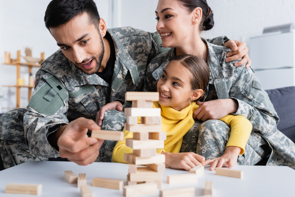 Muž ve vojenské uniformě hraje rozmazané dřevěné bloky hra v blízkosti manželky a dítě doma  - Fotografie, Obrázek