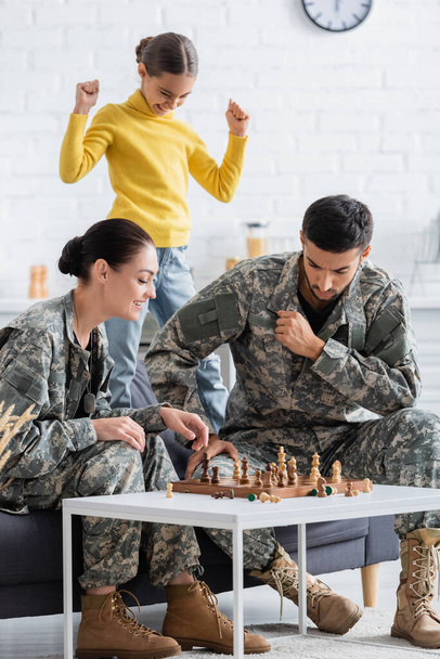 Parents en uniforme militaire jouant aux échecs près de leur fille à la maison  - Photo, image
