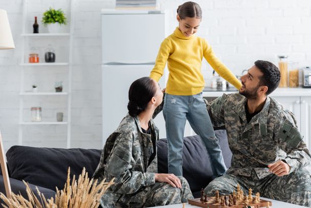 Happy parents in camouflage uniform looking at preteen girl near chess at home  - Photo, Image