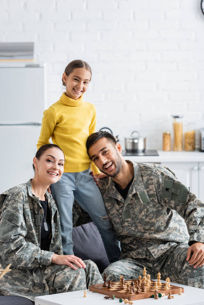 Happy kid standing near parents in military uniform looking at camera near chess at home  - Фото, изображение