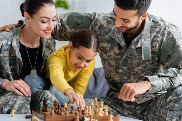 Gülümseyen çocuk, ailelerinin yanında satranç oynuyor. Evde kamuflaj üniformasıyla.  - Fotoğraf, Görsel