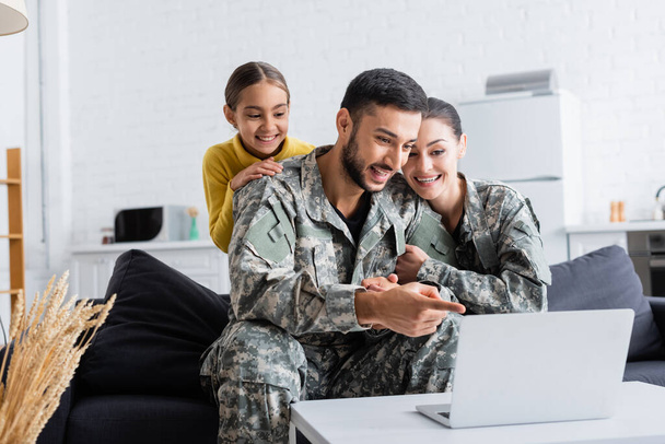 Lächelnder Mann in Tarnuniform zeigt auf Laptop in der Nähe von Frau und Kind zu Hause  - Foto, Bild