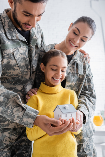 Sonrientes padres en uniforme militar sosteniendo el modelo de casa cerca del niño en casa  - Foto, Imagen