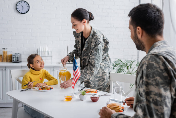 Lächelnde Frau in Militäruniform gießt Orangensaft neben Familie und amerikanischer Flagge auf den heimischen Tisch  - Foto, Bild