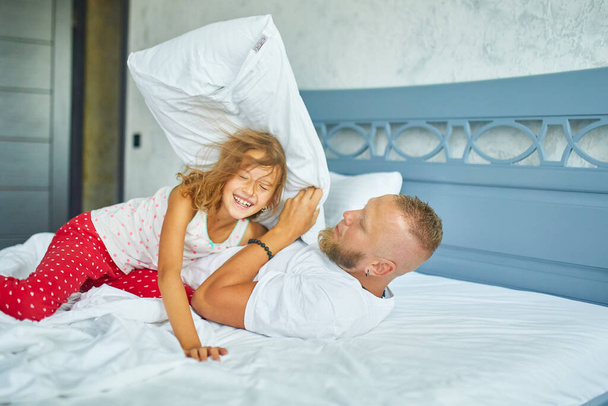 Szczęśliwa córka i ojciec bawiący się w walkę na poduszki w łóżku rano w domu, walcząc z poduszkami, bawiąc się razem w domu w weekend rodzinny - Zdjęcie, obraz