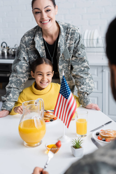 Mujer sonriente en uniforme militar de pie cerca de su hija, desayuno y bandera americana en la cocina  - Foto, Imagen