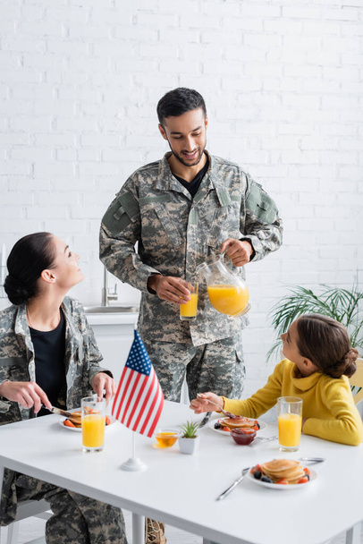 Hombre en uniforme militar vertiendo jugo de naranja cerca de la familia y la bandera americana durante el desayuno en casa  - Foto, Imagen